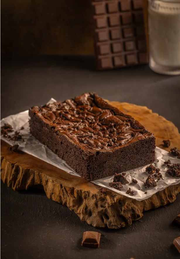 Brownies cokelat utuh di atas papan kayu dengan tekstur padat dan taburan cokelat